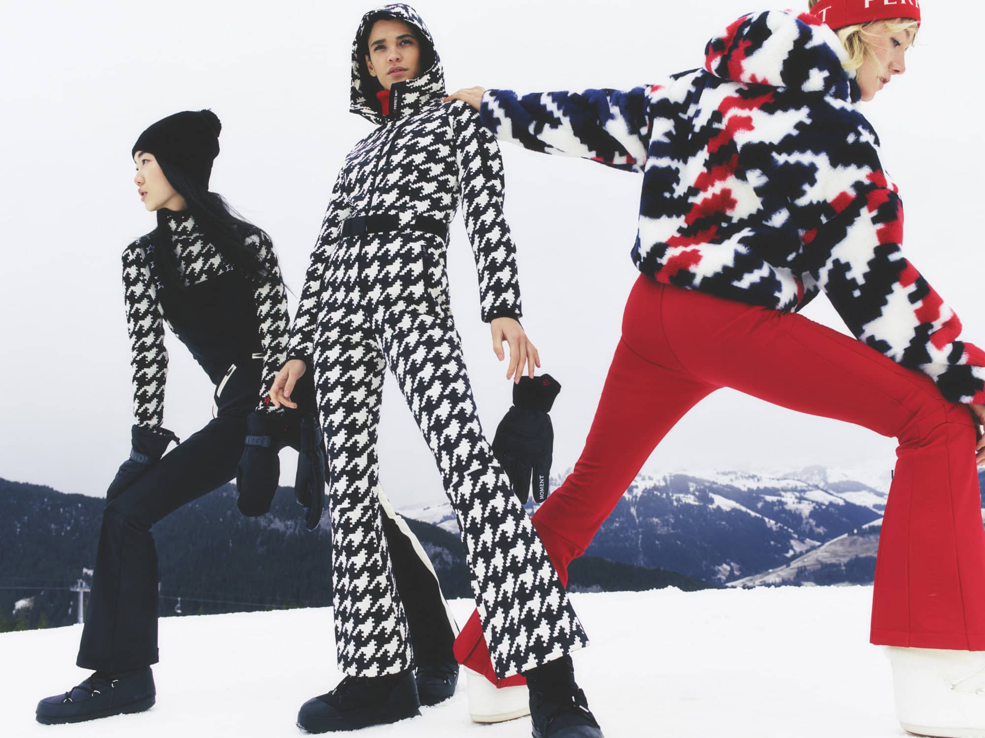 Three fashion models in ski apparel, one in a black houndstooth-patterned jumpsuit, one in a red and white printed jacket and red pants, and one in black.