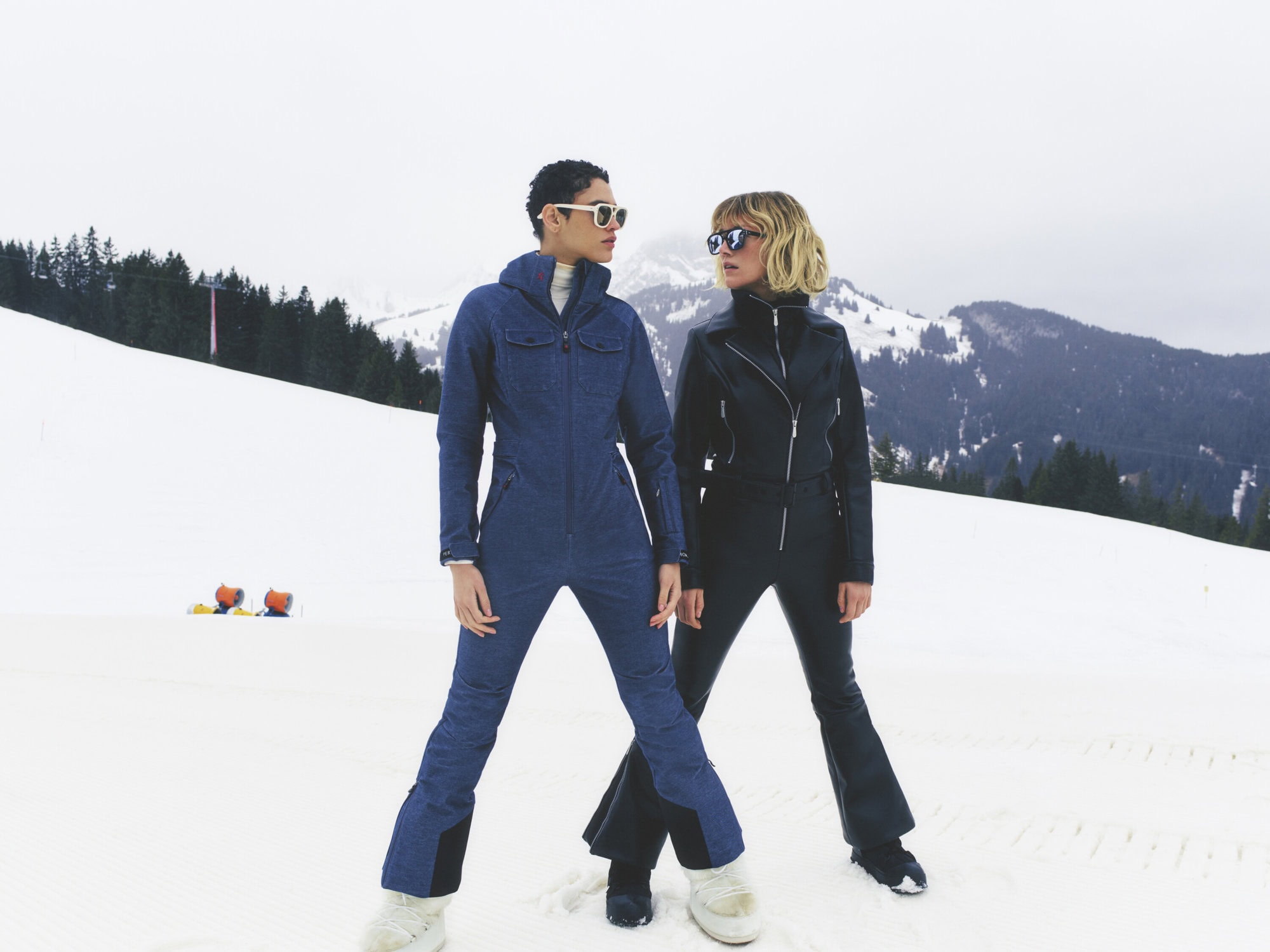 Two women pose in matching ski suits in a snowy mountain landscape. The outfits are blue and black with white snow boots.