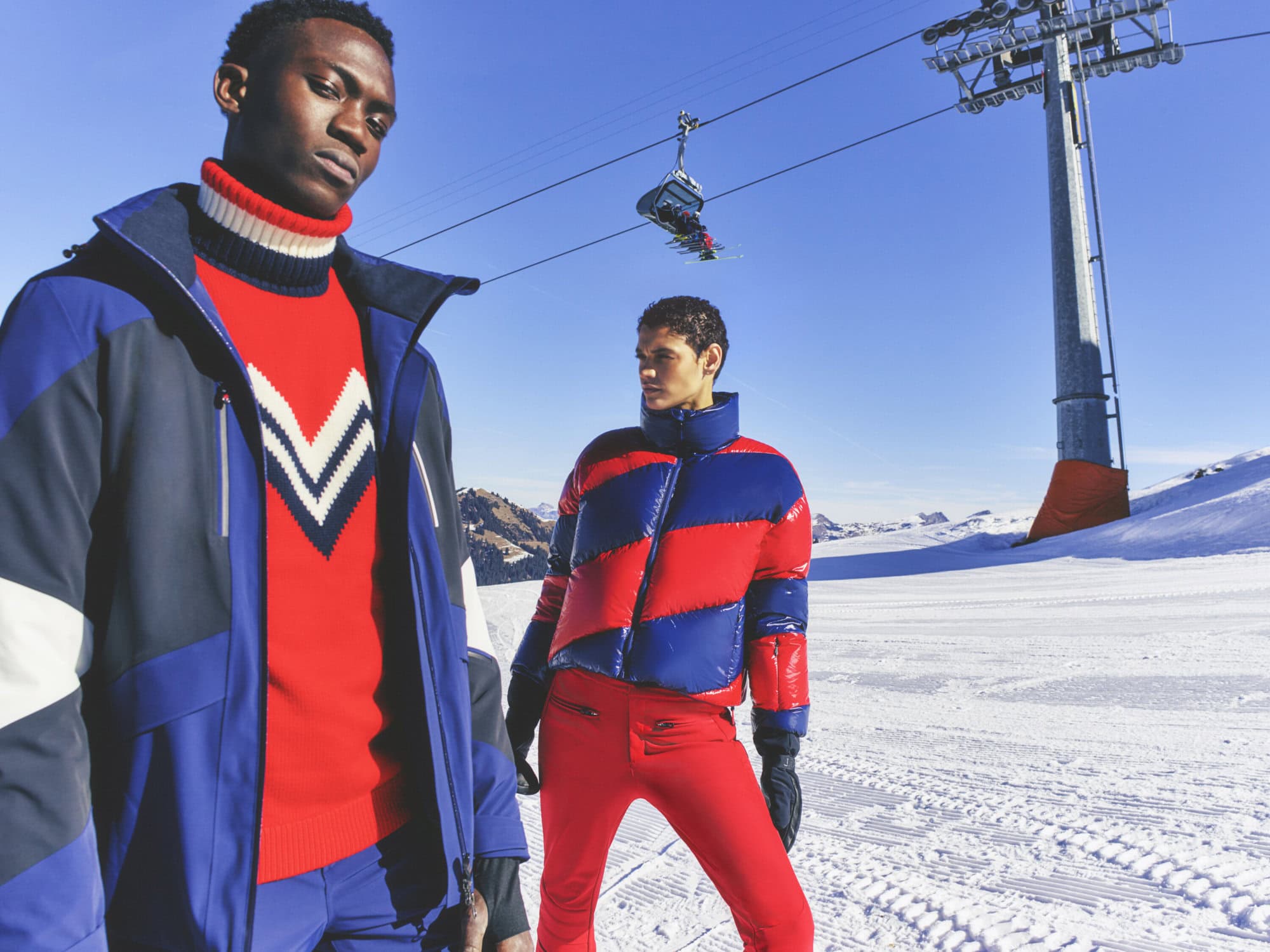 Two adult male models wearing winter ski and snowboard fashion stand against a snowy mountain backdrop under a bright blue sky.
