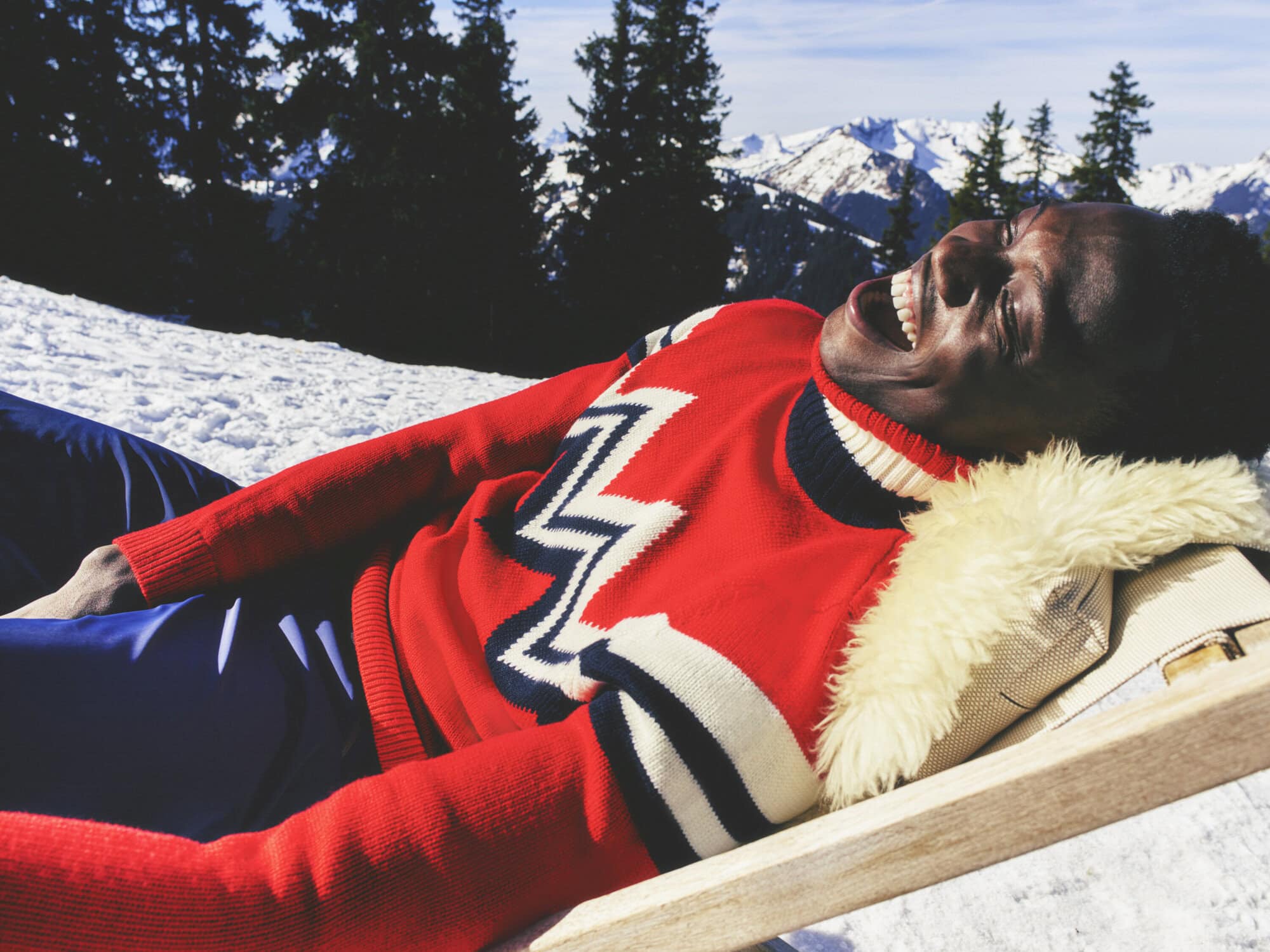 An adult male laying on a lounge chair on a snowy surface wearing a red and white sweater smiling with snow-covered mountains in the background.