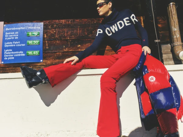 A woman in ski apparel sits casually against a wooden wall outdoors, with a colorful down jacket, ski boots and mountain sign.