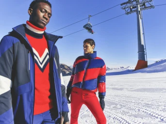 Two models in red and blue ski apparel standing on a snowy mountain with a ski lift in the background.