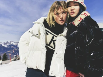 Two women posing outdoors against a snowy mountain landscape wearing winter puffer jackets and branded ski apparel