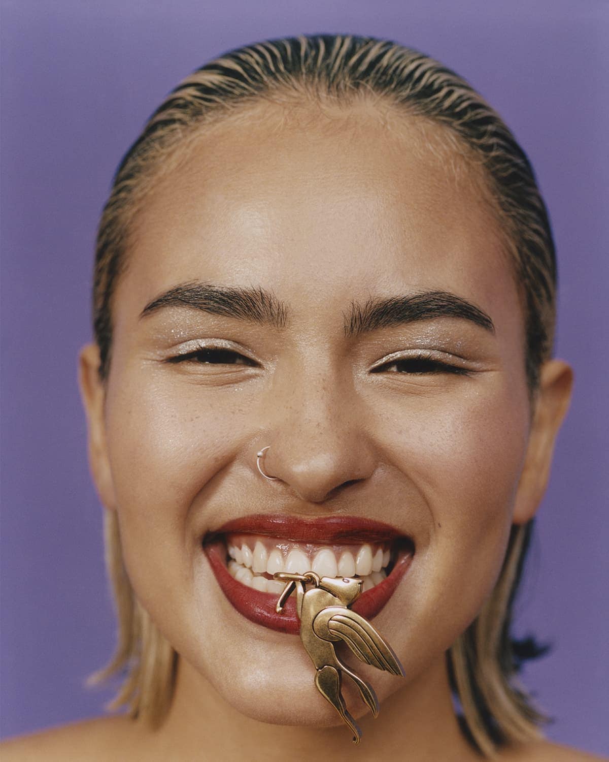 Close-up portrait of a smiling woman with blonde hair, wearing red lipstick, and holding a gold winged figure in her mouth against purple backdrop.