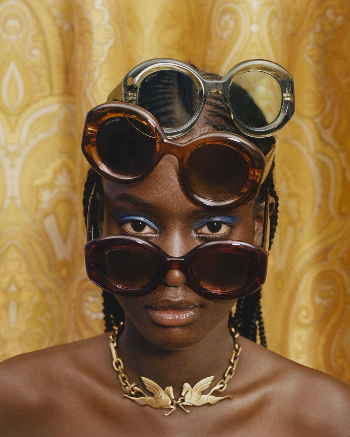 Close-up portrait of a Black woman wearing layered oversized sunglasses and a gold winged necklace against a patterned yellow background.
