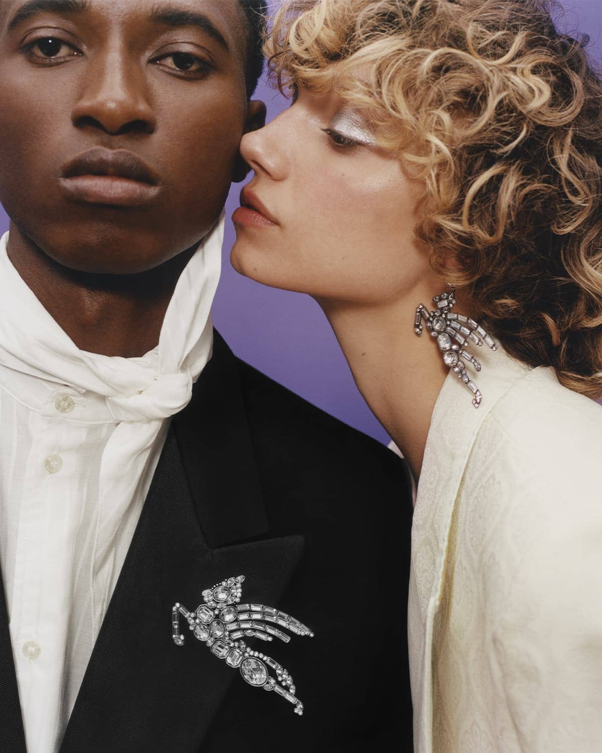Close-up of a stylish couple: an African American male in a black suit, and a Caucasian female with curly hair and silver eyeshadow.