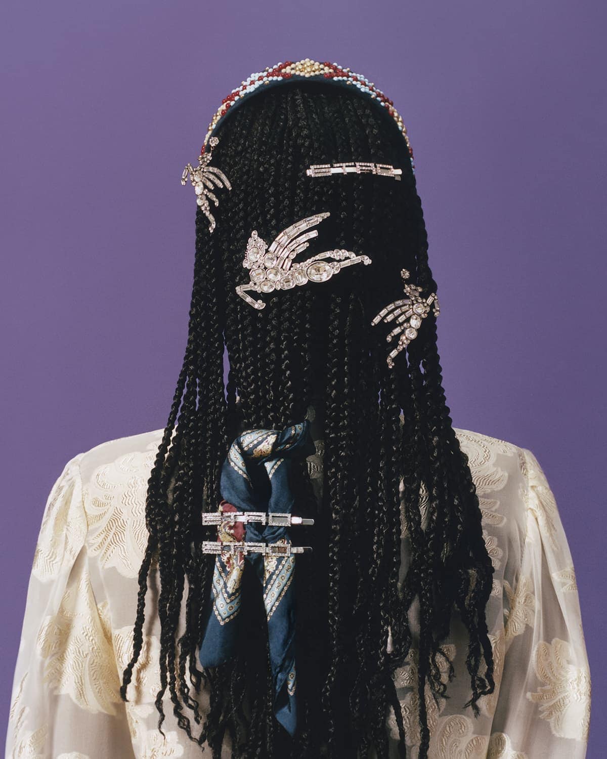 Braided black hair styled with jeweled barrettes and decorative scarf, view from behind on purple background with pale yellow floral top.