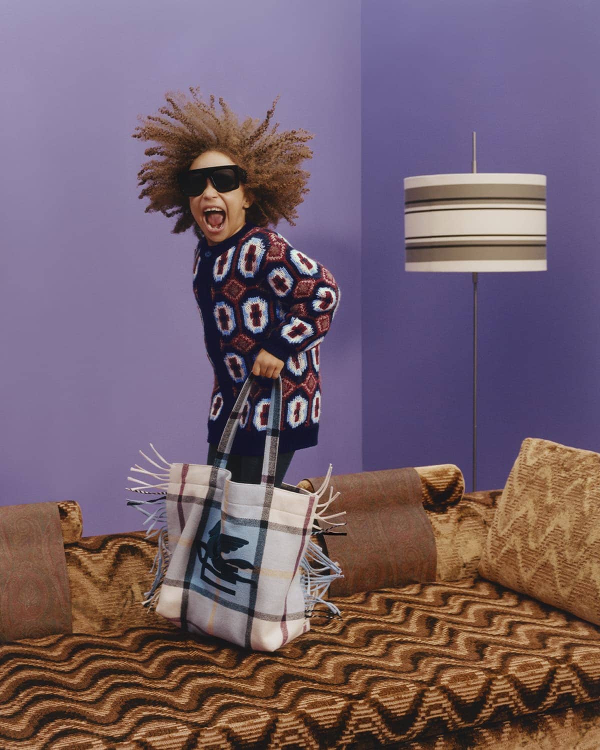 A child with an afro wearing sunglasses and patterned tunic holds a tote bag while posing on a patterned couch against a purple wall.