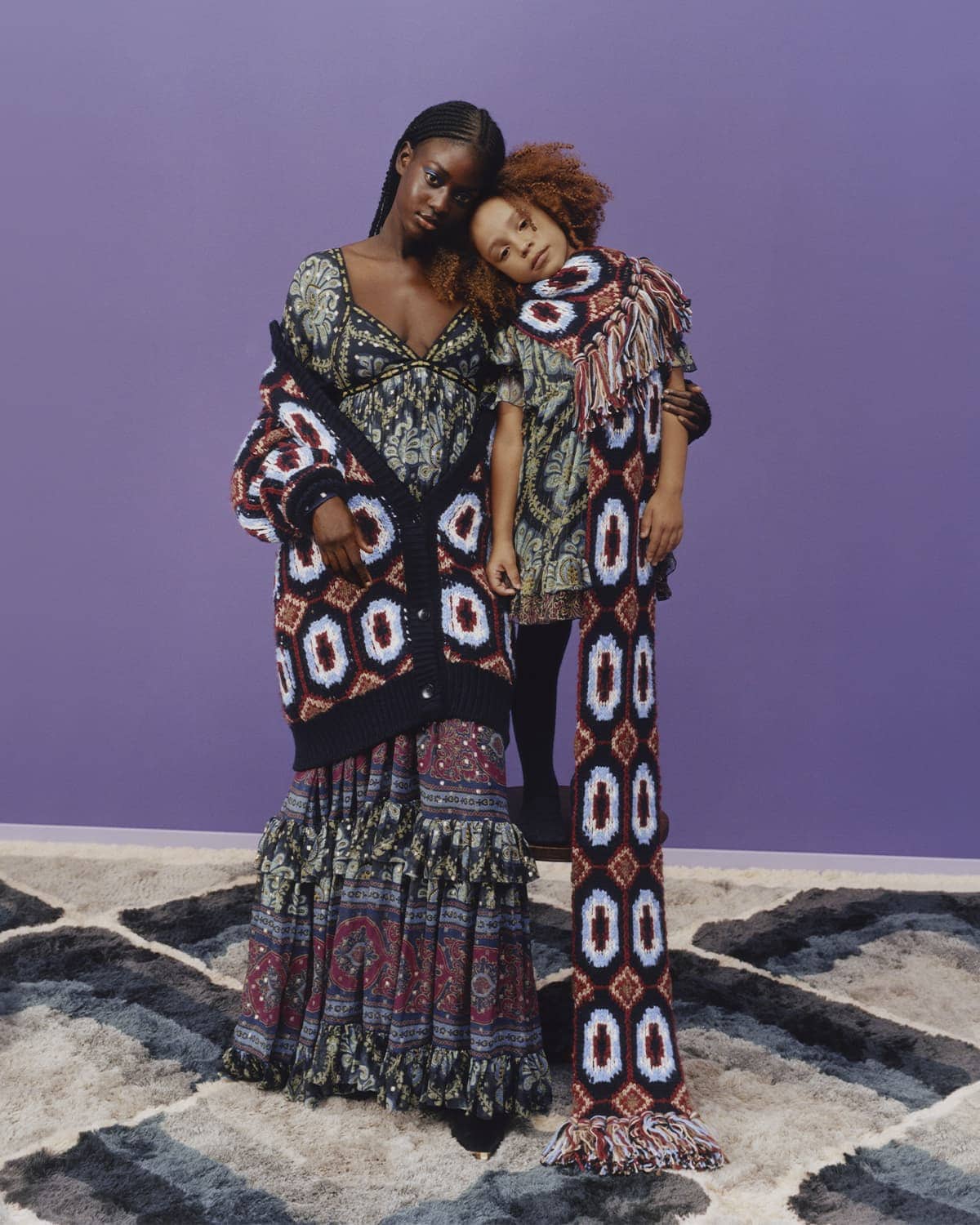 Two women wearing patterned dresses and knitwear stand on a patterned carpet in front of a lavender backdrop, fashion photography.