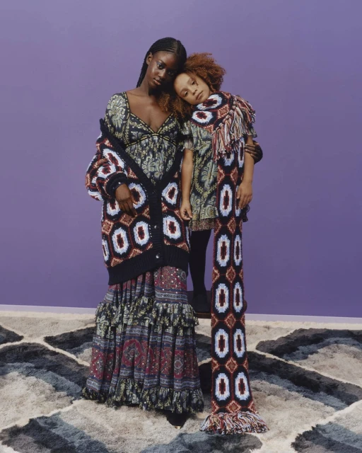 Two women wearing patterned dresses and knitwear stand on a patterned carpet in front of a lavender backdrop, fashion photography.