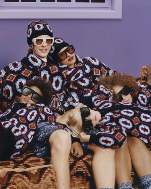 Group of five people wearing matching patterned sweaters and sunglasses reclining on a brown patterned couch against a purple wall.