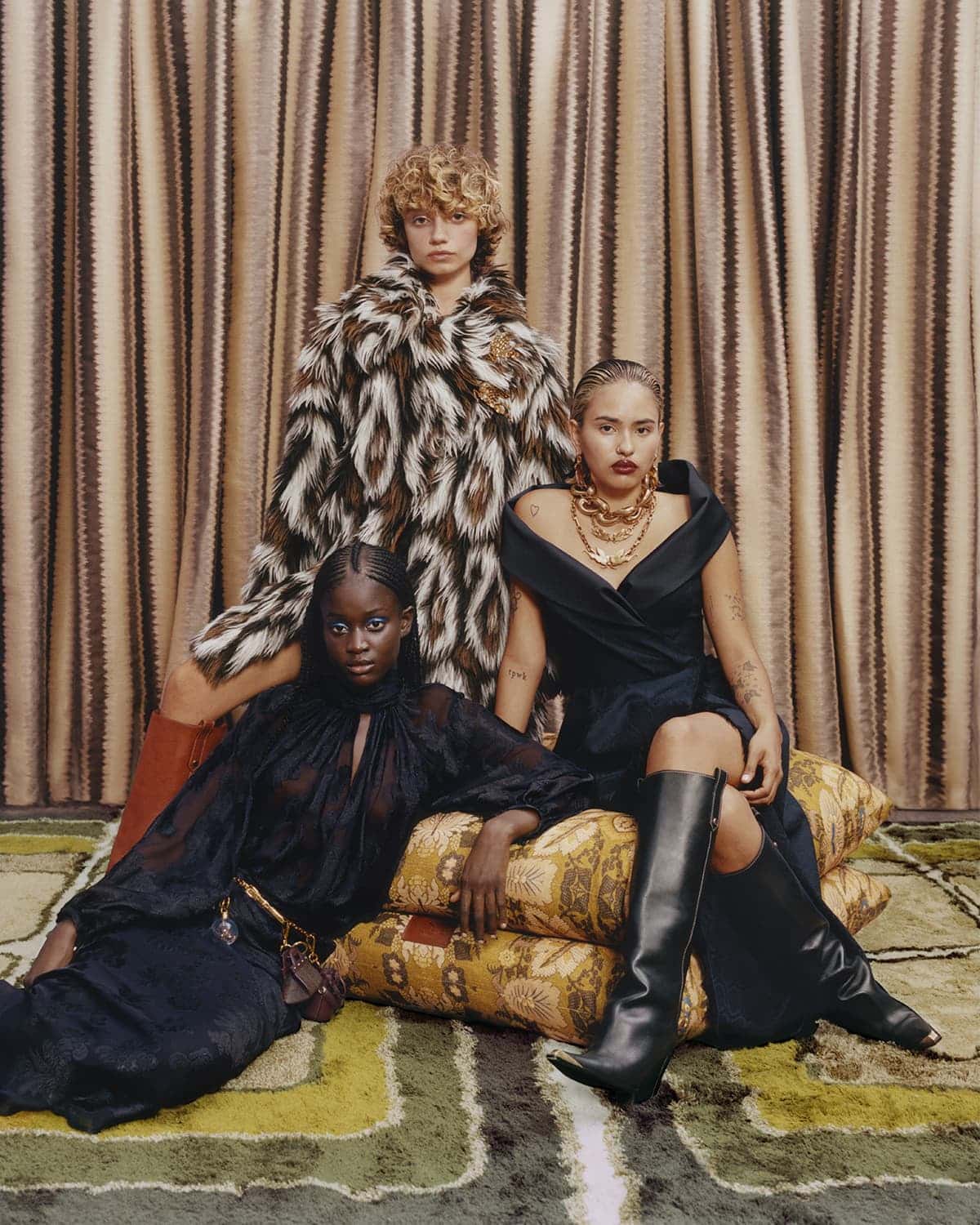 Three fashion models pose together indoors. Two are sitting on patterned pillows while another stands behind them in a patterned coat.