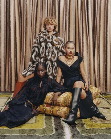 Three fashion models pose together indoors. Two are sitting on patterned pillows while another stands behind them in a patterned coat.