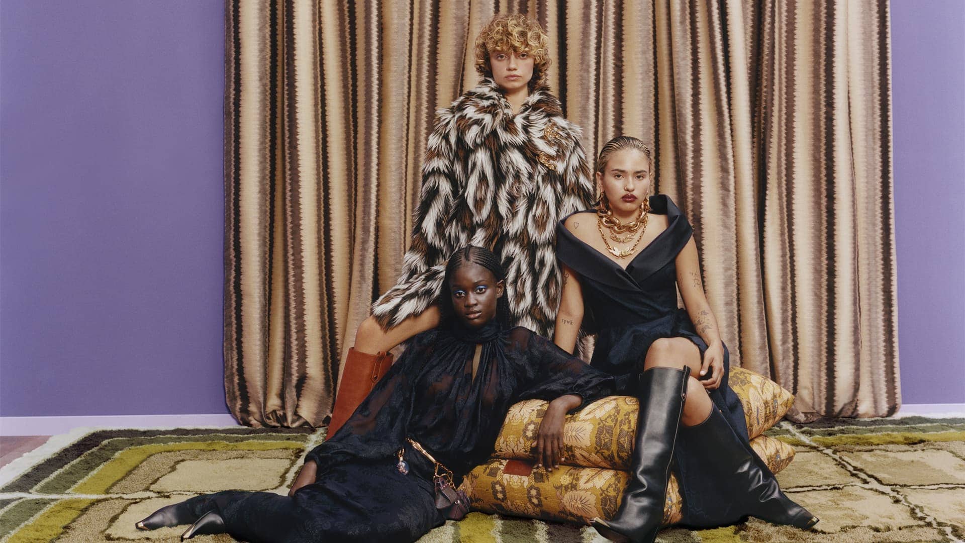 Three fashion models in a photo studio. Two seated on patterned pillows wearing black dresses and knee-high boots, one standing in fur coat.
