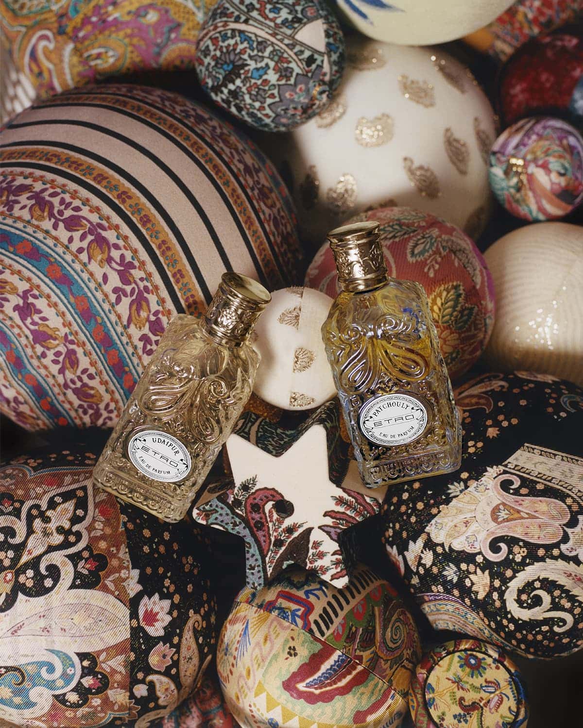 Two glass perfume bottles with golden caps are surrounded by a mix of colorful patterned textile spheres and a white star-shaped object.