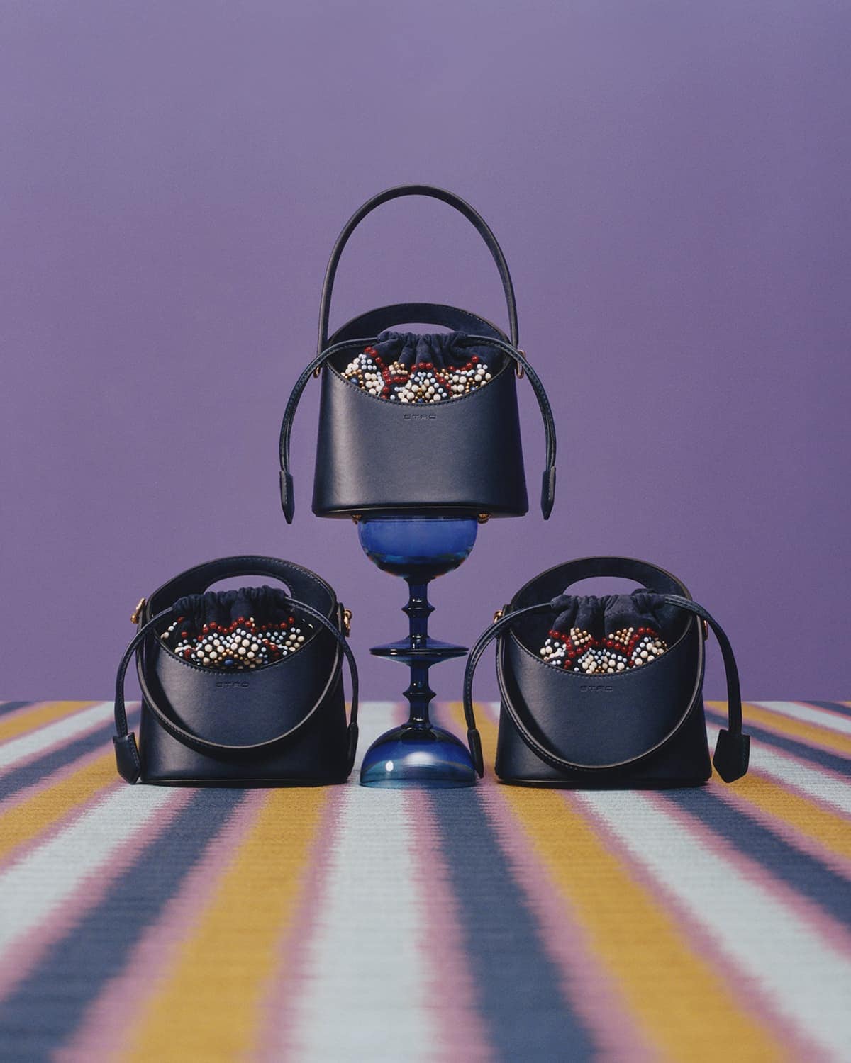 Three leather bucket bags with handles, filled with beaded drawstring closures, styled on a blue glass pedestal and striped surface.