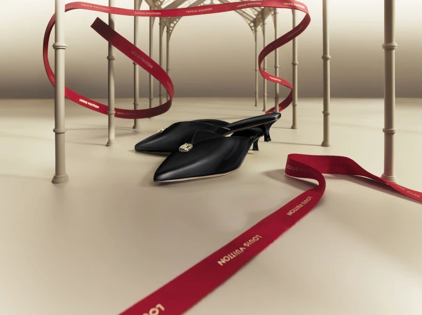 Black mule heels with gem embellishments set against a light backdrop with floating red logo ribbon accents and archway structures.