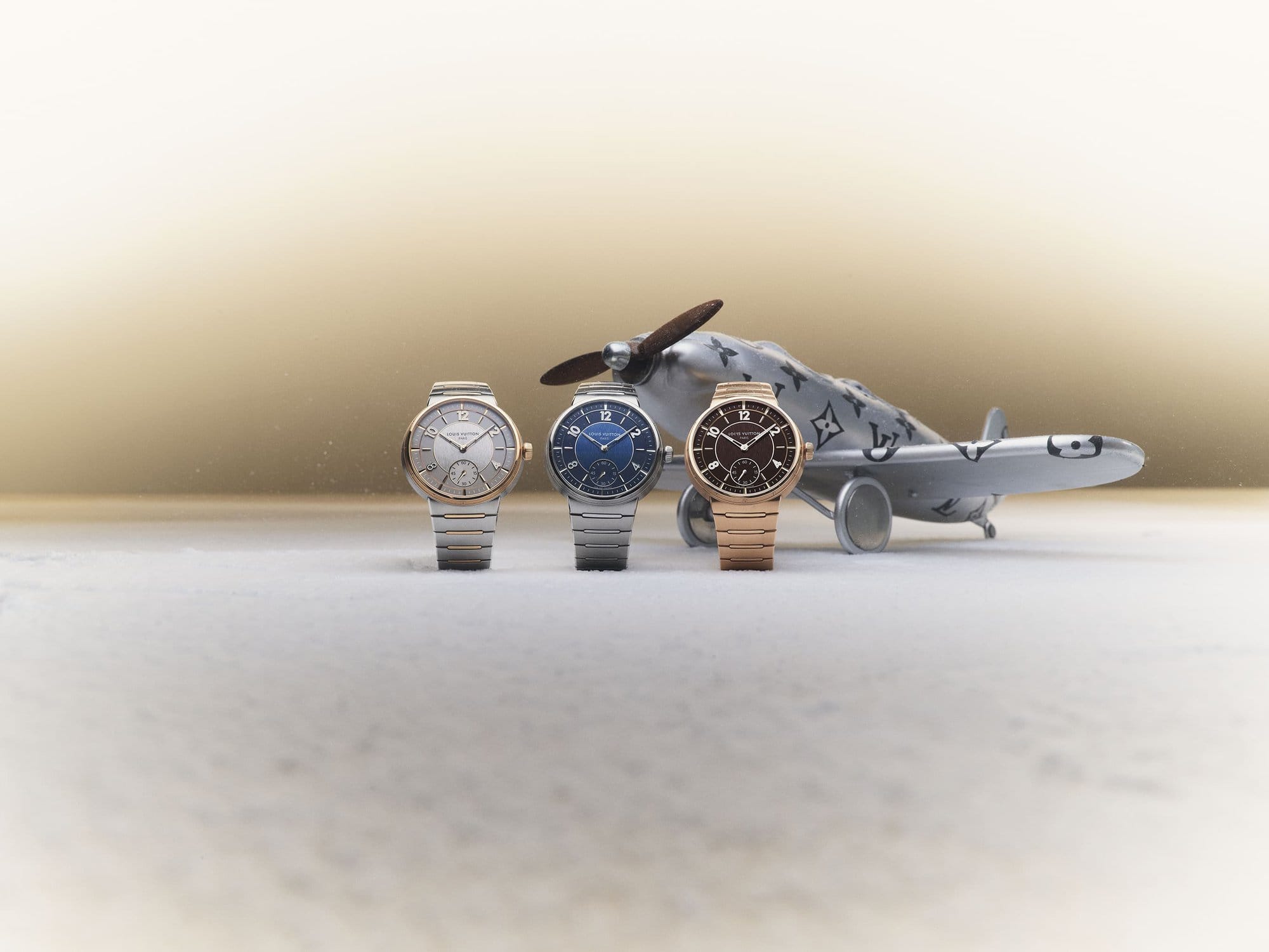 Trio of luxury wristwatches, featuring gold, silver, and blue dials, displayed in front of a Louis Vuitton airplane toy.