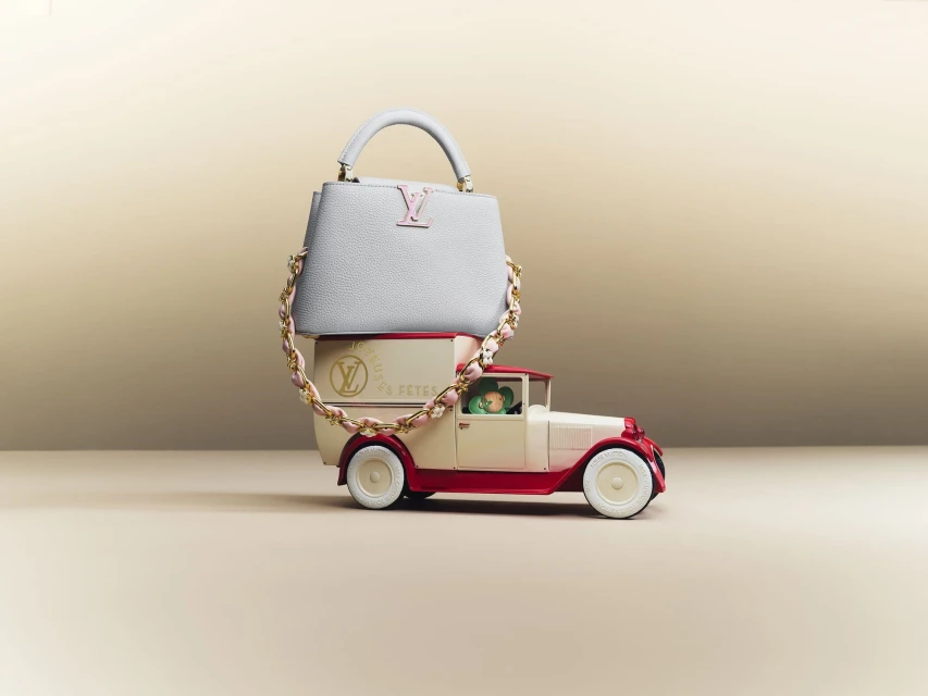 Light blue Louis Vuitton Capucines Mini handbag with pink and gold chain atop a vintage cream and red toy truck against a light tan backdrop.