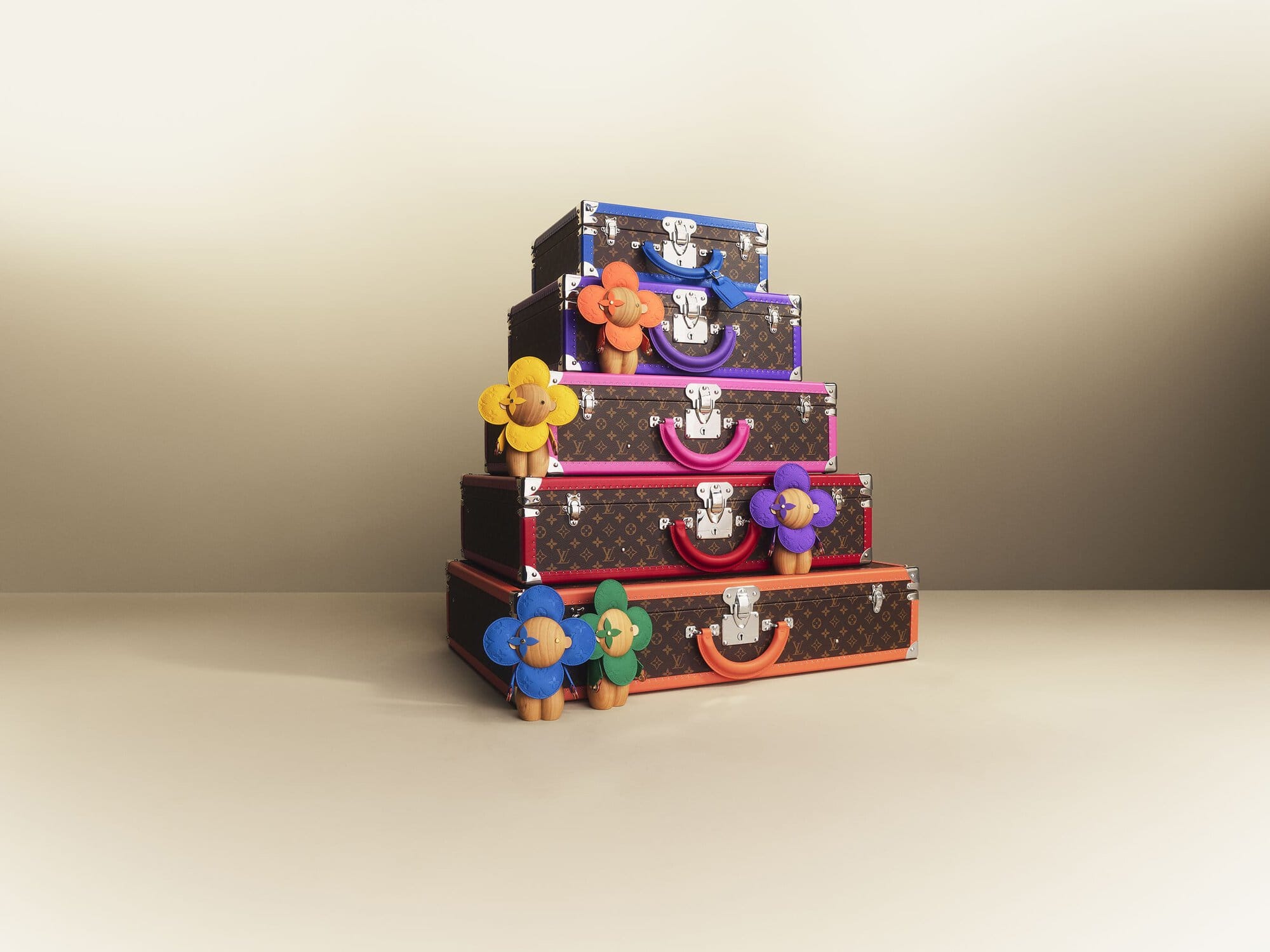 Stack of Louis Vuitton Boite Flacons vanity cases with bright trim and flower figures on a beige backdrop, showcasing luxury travel accessories.