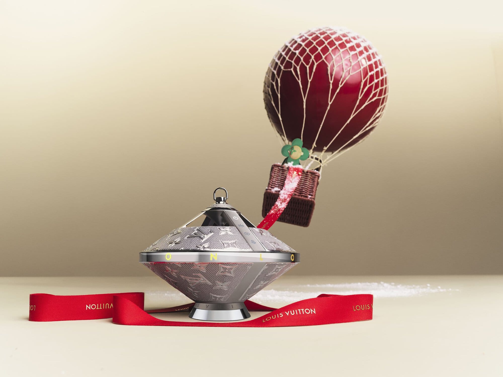 Still life of a Louis Vuitton ornament featuring a silver-toned base and a hot air balloon topper against a neutral background with red ribbon.