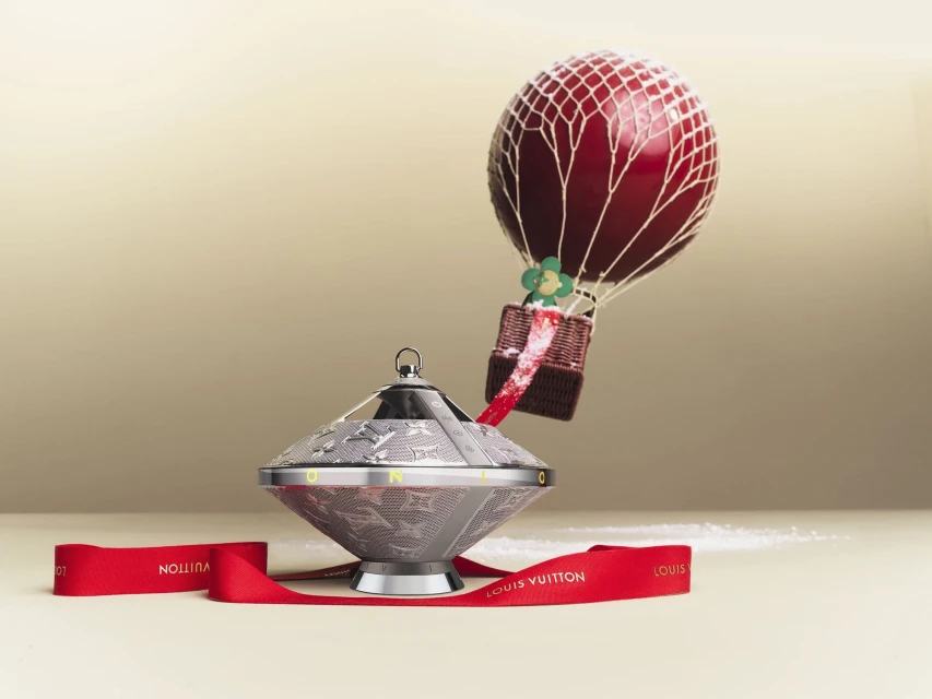 Still life of a Louis Vuitton ornament featuring a silver-toned base and a hot air balloon topper against a neutral background with red ribbon.