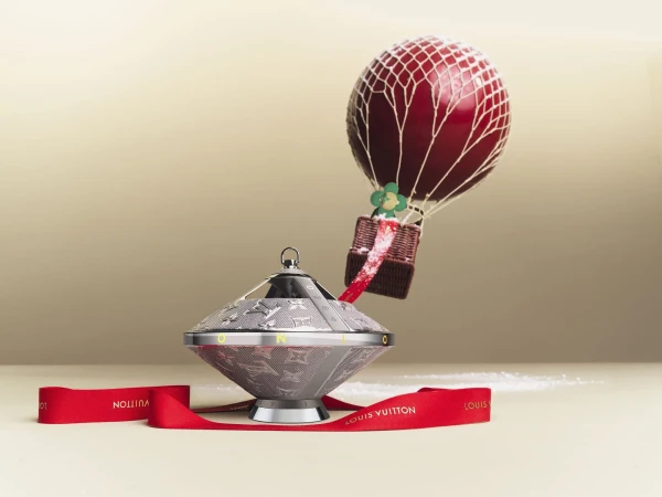 Still life of a Louis Vuitton ornament featuring a silver-toned base and a hot air balloon topper against a neutral background with red ribbon.