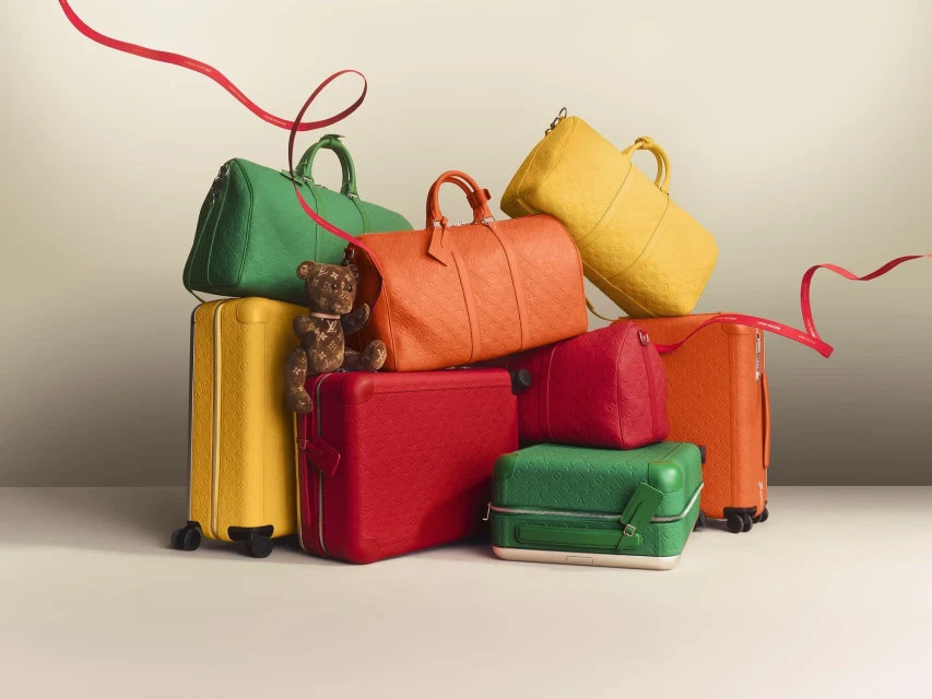 Pile of colourful leather luggage and bags, including rolling suitcases and duffel bags, with a brown teddy bear, and a red ribbon.