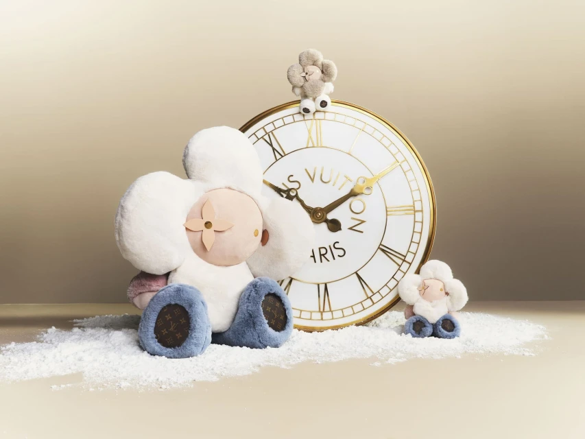 Louis Vuitton stuffed flower dolls sit alongside a clock on a white surface, showcasing luxury fashion and plush toy aesthetics.
