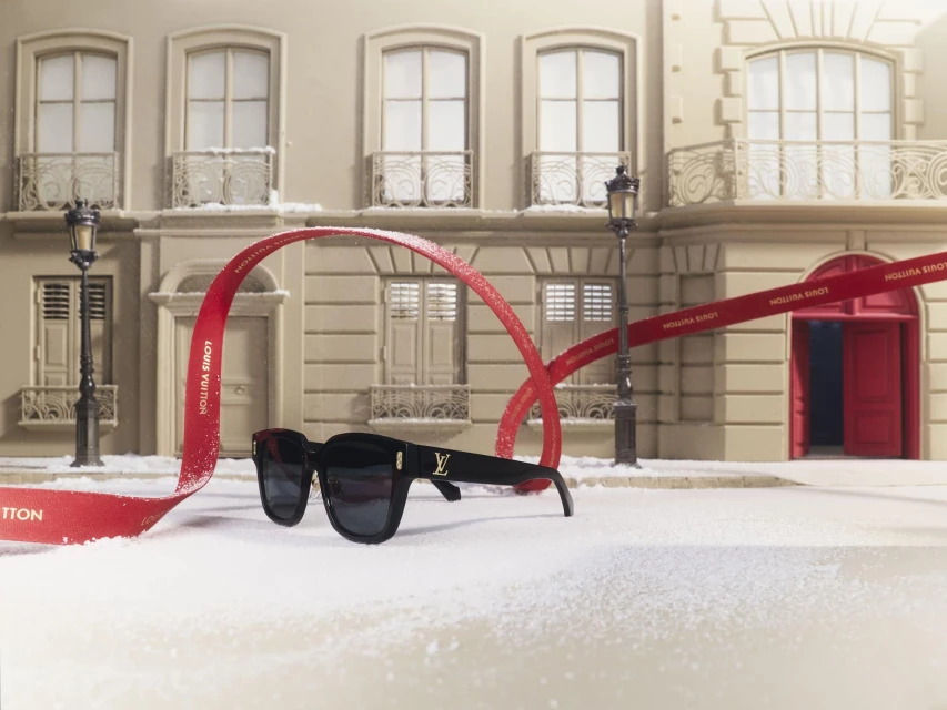 Louis Vuitton sunglasses on snowy surface with red ribbon, against a building backdrop, highlighting luxury fashion accessories.