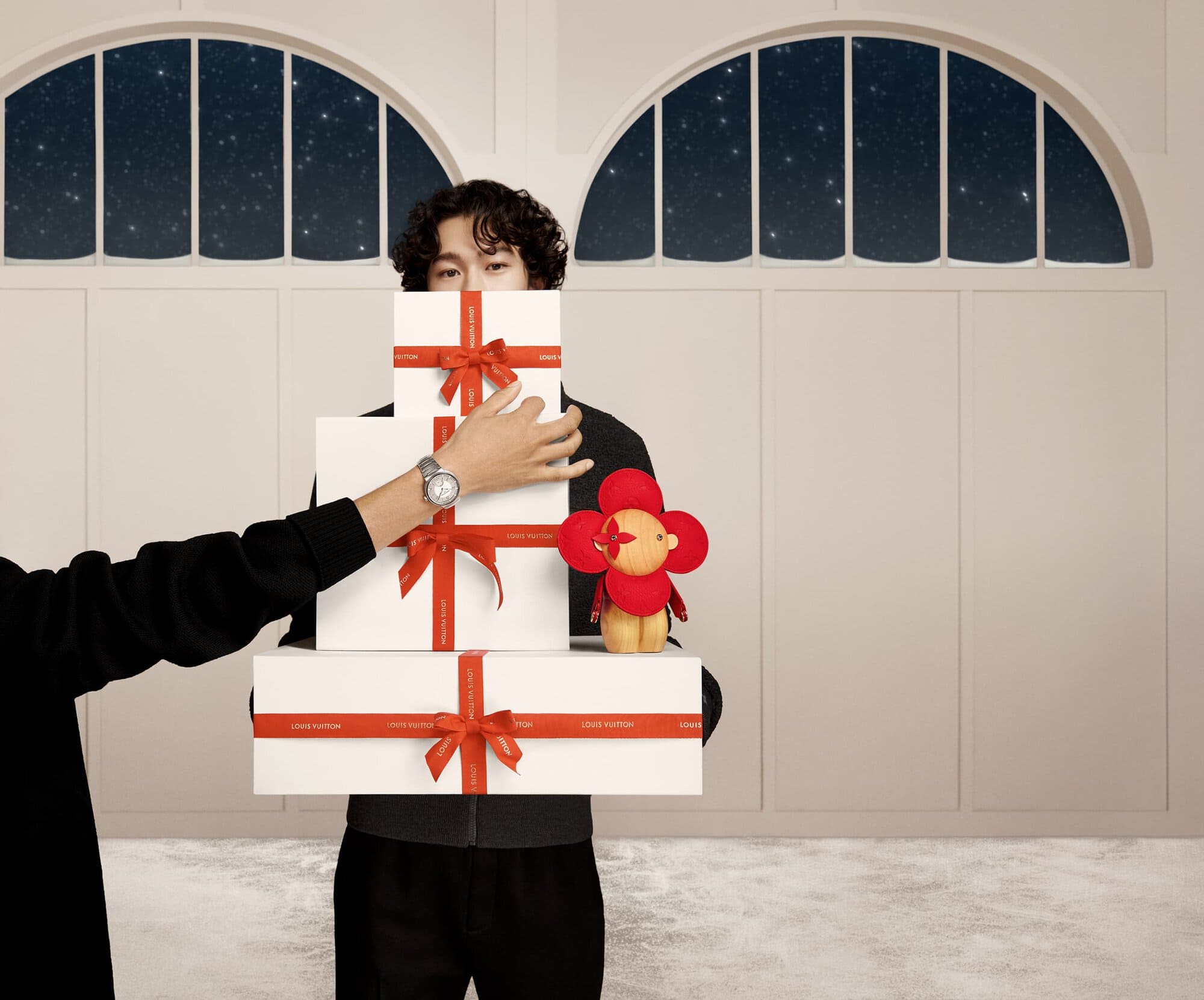 Person holding a stack of gift boxes with red ribbons and a flower doll in front of a white panel wall and arched window.