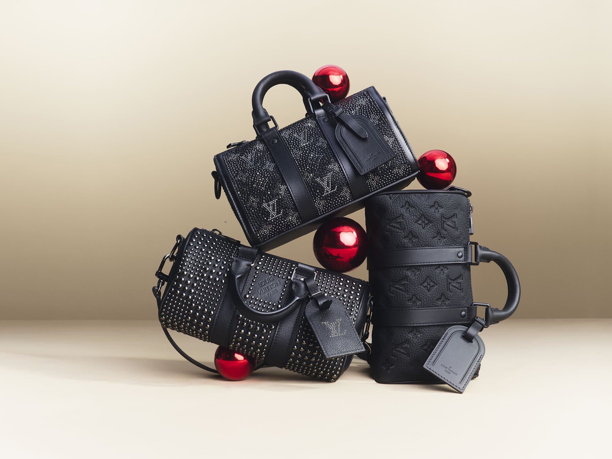 Three black Louis Vuitton bags are stacked with three red ornaments against a gradient tan background in a product studio shot.