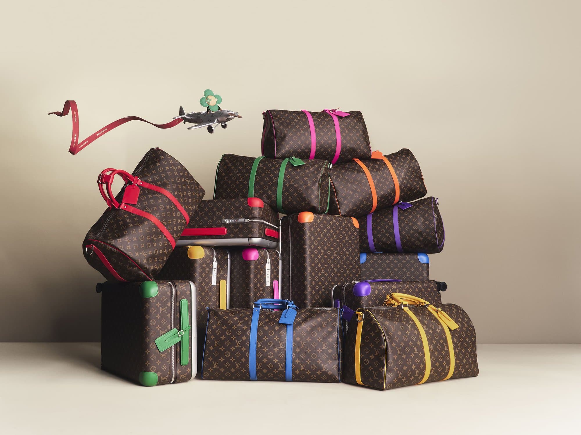 Stack of Louis Vuitton luggage with multicolored straps, a small airplane with a red ribbon flies above the pile against a neutral background.
