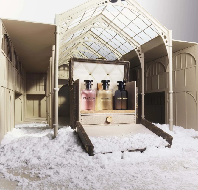 Three perfume bottles inside a Louis Vuitton trunk displayed in an architectural model of a greenhouse with white snowy ground and neutral tones.