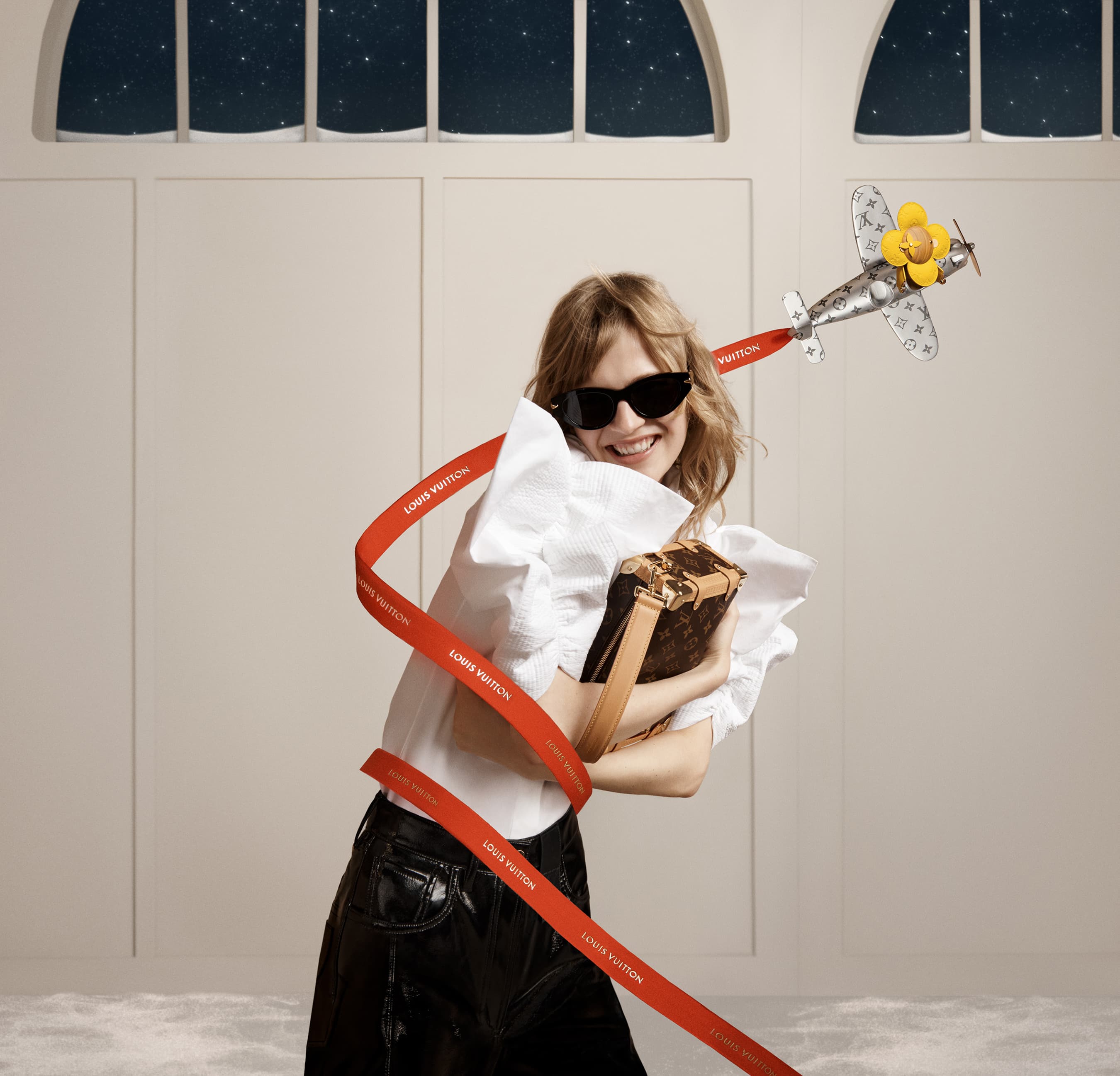 A smiling woman in sunglasses holds a box handbag next to a toy airplane with a red ribbon indoors. A window with a starry scene appears.