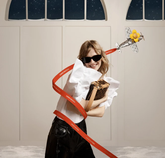 A smiling woman in sunglasses holds a box handbag next to a toy airplane with a red ribbon indoors. A window with a starry scene appears.