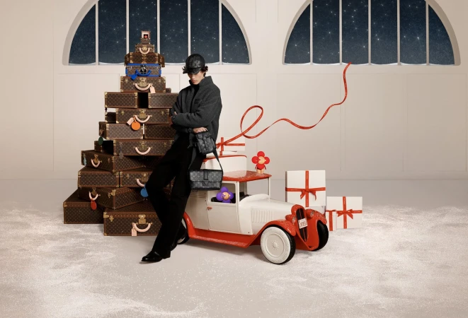 A man poses in front of a stack of luggage and a vintage toy car in a whimsical studio scene with gift boxes, set against a starry backdrop.