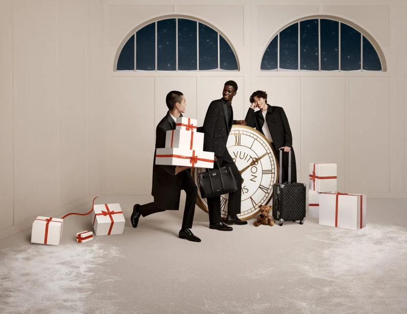 Three people dressed in formal attire are gathered around a large clock and a collection of gift boxes in a bright indoor setting.
