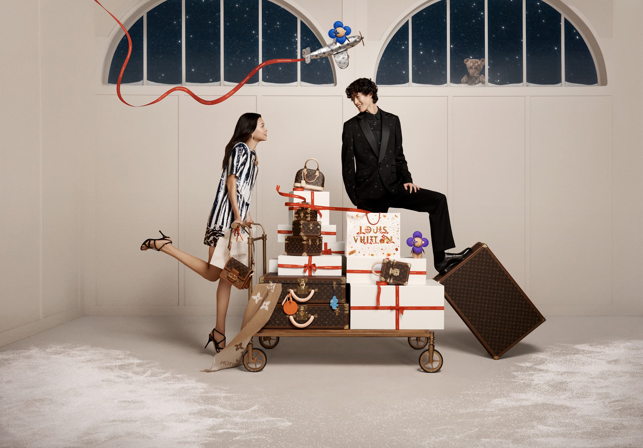 Two fashion models pose in a studio with Louis Vuitton luggage stacked on a luggage cart, a toy airplane, and winter-themed decorations.