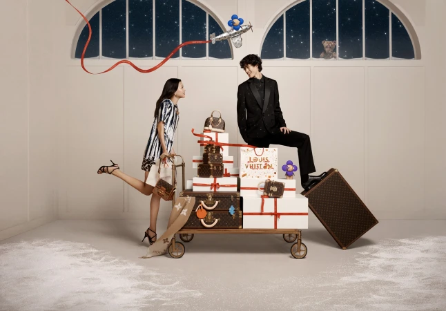 Two fashion models pose in a studio with Louis Vuitton luggage stacked on a luggage cart, a toy airplane, and winter-themed decorations.