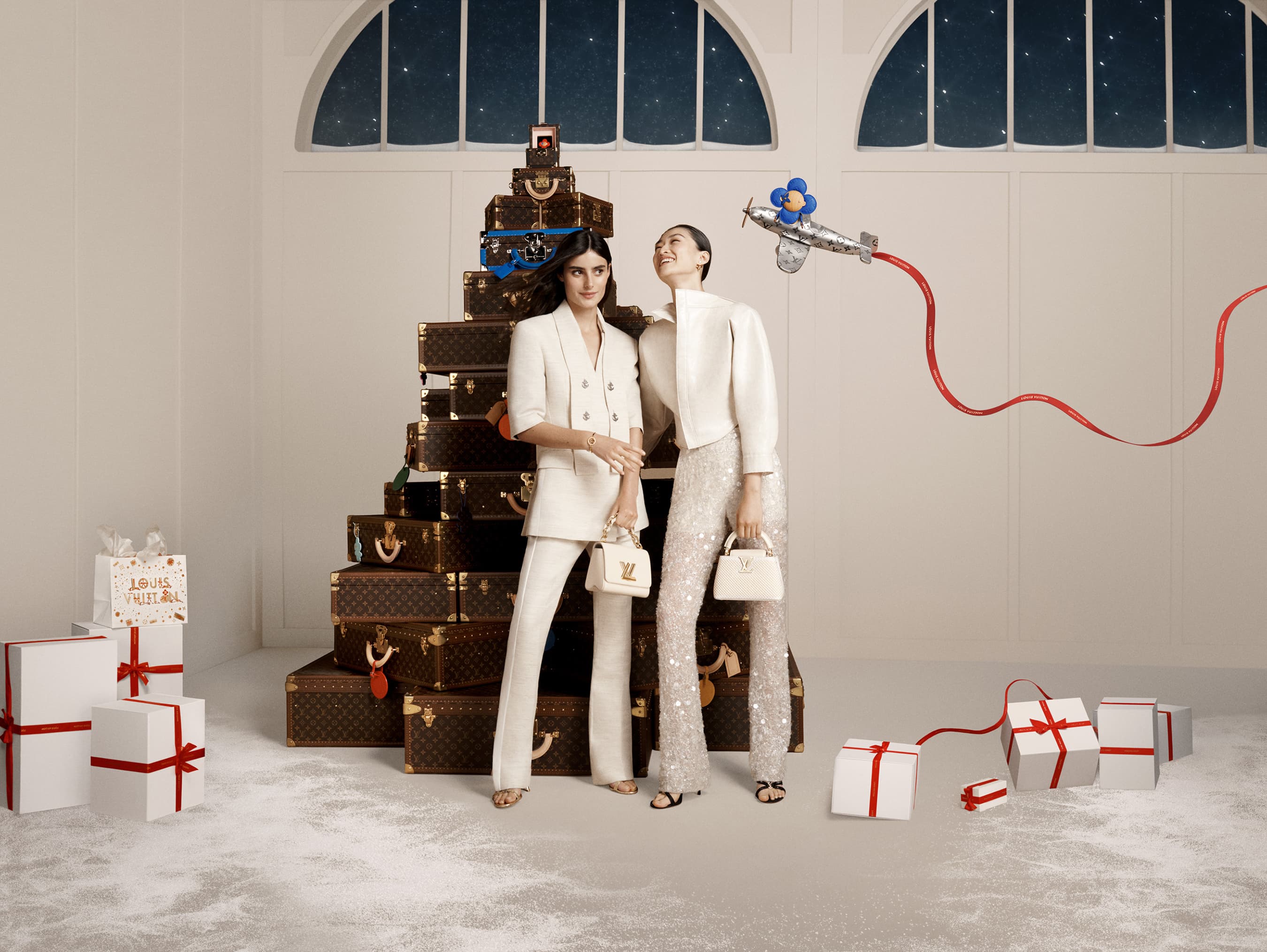Two women pose in a holiday fashion shoot with Louis Vuitton luggage stacked as a Christmas tree, surrounded by gift boxes.