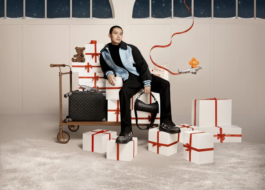 Stylish man in black suit and blue jacket sits atop wrapped gift boxes, holding purse, with luggage and toy airplane and bear.