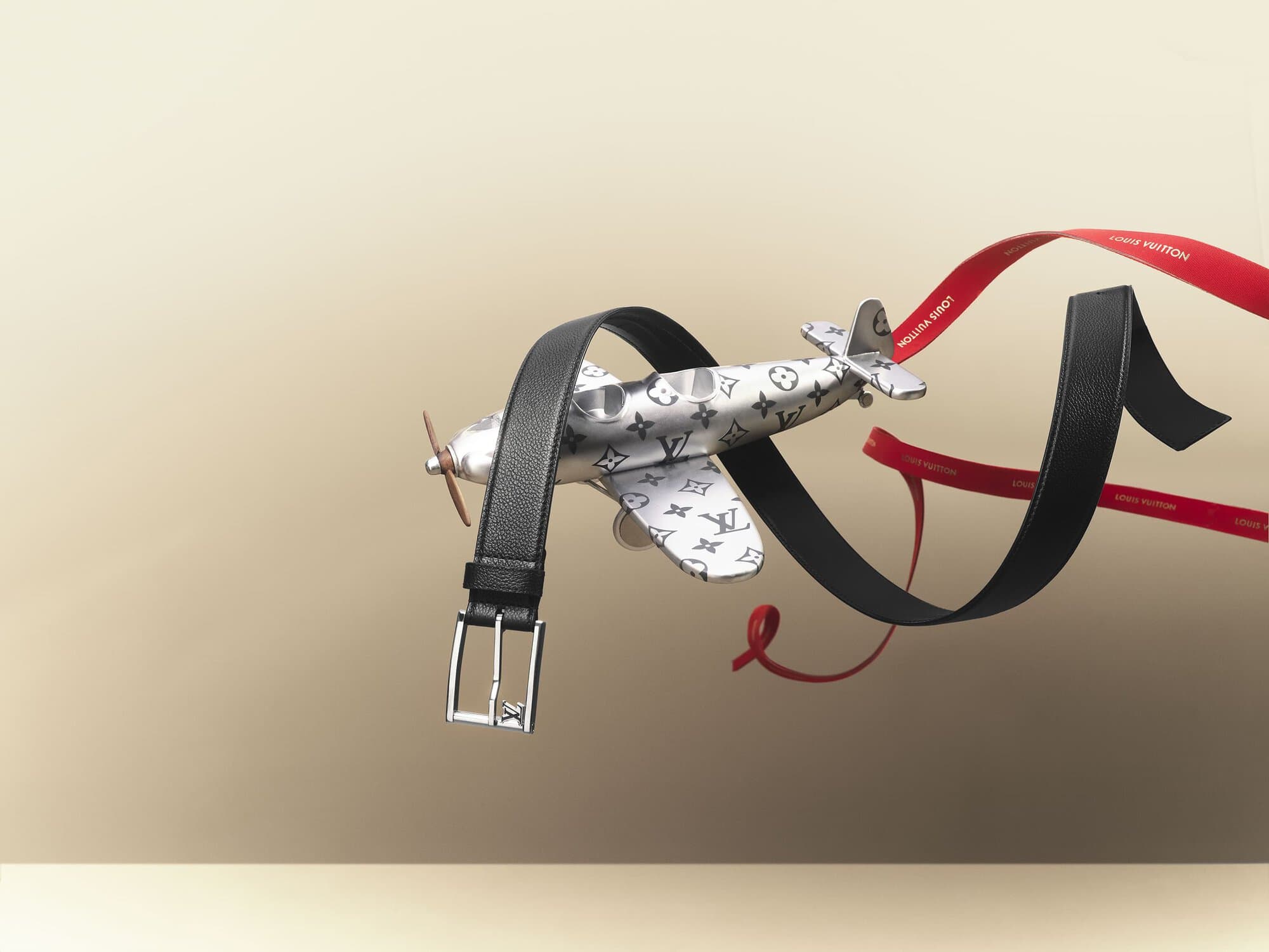 Luxury black leather belt with silver buckle levitates with a toy airplane featuring a Louis Vuitton pattern on a beige gradient background.