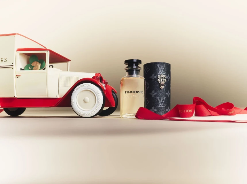 Still life featuring Louis Vuitton perfume bottle and cylindrical case beside a toy truck with a teddy bear and red ribbon.
