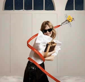 Smiling woman in dark sunglasses and white ruffled blouse holding a monogram handbag wrapped in a red ribbon for Louis Vuitton