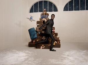 Black man in a black suit carrying a teal duffle bag past a tall stack of monogrammed luggage trunks.