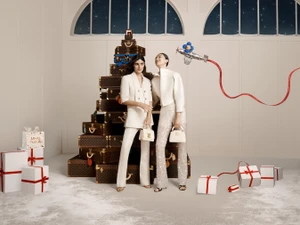 Two female models posing in white outfits in front of a tall stack of monogrammed Louis Vuitton trunks.