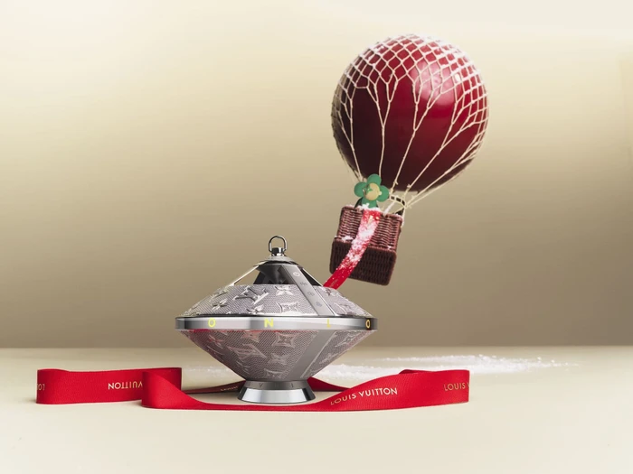 Monogram silver speaker tethered to a red hot air balloon with white netting on a neutral background for Louis Vuitton Holiday 2023