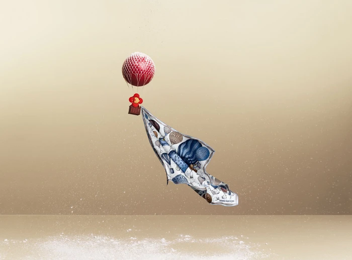 Red hot air balloon carrying a patterned silk scarf against a gradient beige studio background