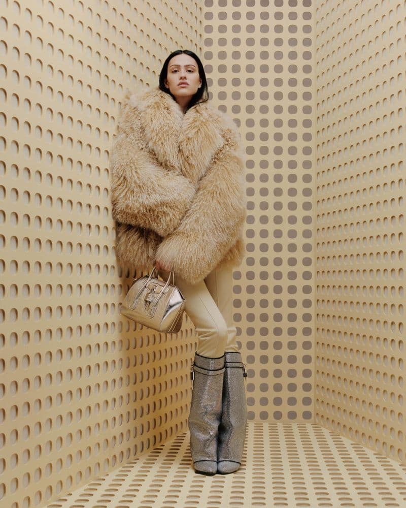 Fashionable woman in fur coat, pants, and shiny boots poses in studio with patterned walls and floor, holding a handbag for a stylish look.